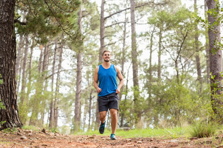 Male runner in blue sportswear jogging along dirt trail through dense pine forest under sunlight....