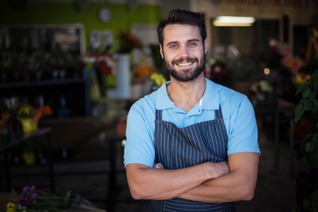 Bearded man crossing arms, smiling inside floral shop wearing striped apron among vases and plants. Floristry, botanist, artisanal, craft, gardening, fresh, vibrantの写真素材