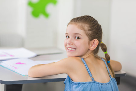 Girl student sitting at gray desk flipping pages in pink heart workbook in classroom, copy space....