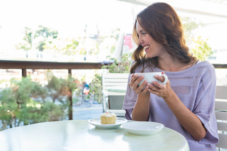Smiling woman in lavender top sitting at outdoor cafÃ© holding coffee mug with cupcake, copy space. Casual, leisure, relaxation, outdoor, vibrant, nature, lifestyleの写真素材