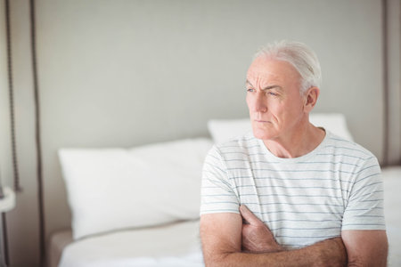 Senior man sitting on bed edge crossing arms gazing pensively over sheets in bedroom, copy space. Aging, contemplation, comfort, serenity, interior, relaxation, homeの写真素材