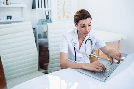 Mid adult medical professional wearing stethoscope and using laptop in exam room, copy space. Healthcare, professionalism, clinical, consultation, modern, sterile, wellnessの写真素材