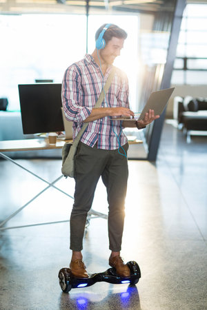 Man riding hoverboard typing on laptop in bright office wearing blue headphones and messenger bag....