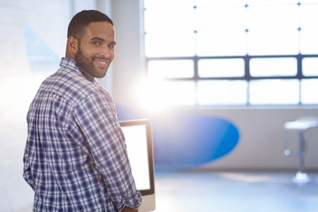 African American man standing at desk in loft office, looking over shoulder with computer monitor....