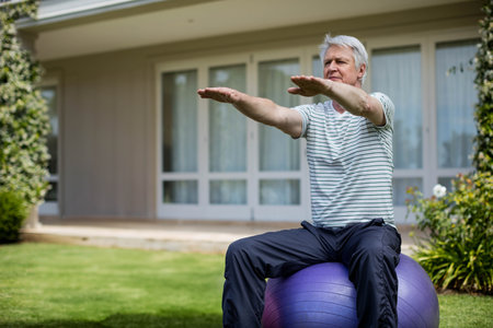 Senior male balancing on purple stability ball on lawn outside home in workout clothes, copy space. Fitness, wellness, outdoor, leisure, relaxation, vitality, activeの写真素材