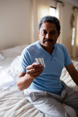 Senior man holding blister pack containing tablets while sitting on bed in bedroom wearing pajamas. Geriatric, healthcare, wellness, relaxation, home, comfort, lifestyleの写真素材
