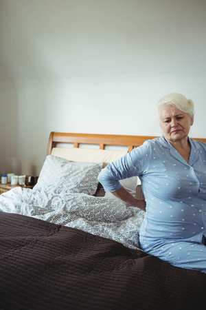 Senior woman sitting on bed in bedroom wearing pajamas supporting back by nightstand, copy space. Comfort, relaxation, wellness, home, pastel, cozy, healthの写真素材
