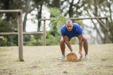 African American man in workout gear lifting wooden log on grass field near rail fence. Fitness, outdoor training, strength, obstacle course, athletic, nature, enduranceの写真素材