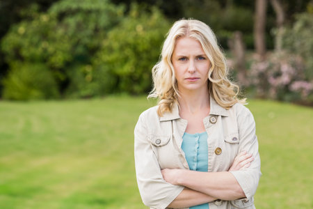 Woman standing with crossed arms wearing beige jacket on lawn in garden surrounded by shrubs. Outdoor, nature, relaxed, tranquility, elegance, foliage, casualの写真素材