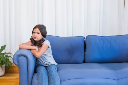 Female child sitting on blue couch in living room with potted plant, white curtains, copy space. Youth, relaxation, domestic, minimalist, comfort, home decor, casualの写真素材