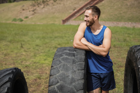Fit man in sportswear leaning with arms crossed on tractor tire at outdoor gym, copy space. Athlete, strength, resilience, rugged, motion, vitality, outdoorの写真素材