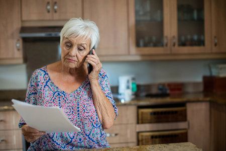 Senior woman talking on smartphone while reading paper in home kitchen near coffee maker. Retirement, communication, domestic, lifestyle, modern, cozy, wellnessの写真素材