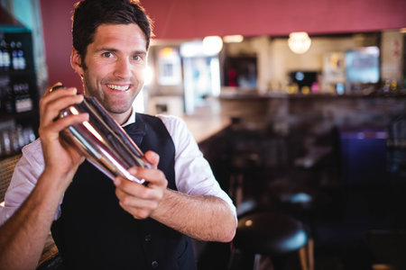 White mid adult male bartender shaking stainless steel cocktail shaker behind wooden bar counter. Bartender, mixology, craft, hospitality, warm lighting, cozy, barwareの写真素材