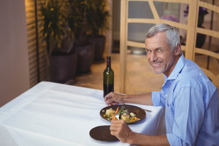 Senior man holding fork and knife at restaurant table with plated meal and wine, copy space. Dining, sophistication, leisure, elegance, ambiance, nutrition, celebrationの写真素材