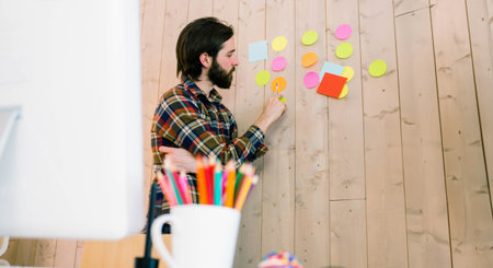 Man placing colorful round and square sticky notes on wooden accent wall in modern office workspace....
