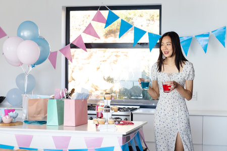 Asian female in floral dress holding red and blue cups in kitchen with balloons, copy space....