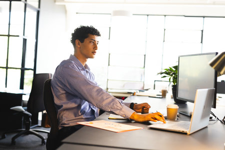 Man in his 30s typing on laptop in modern office with monitor, coffee cup, copy space. Workspace,...