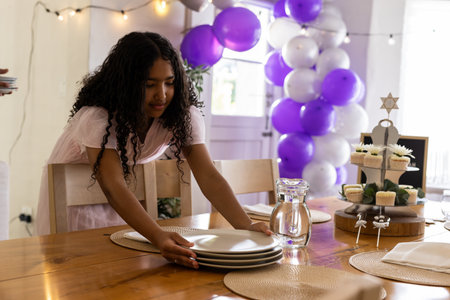Girl leaning at dining table placing stack of plates among cupcakes and balloon arch, copy space. Celebration, party, domestictableware, decor, cheerful, cozy, festiveの写真素材