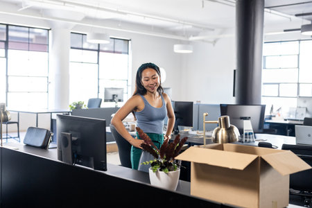 Asian Chinese woman standing unpacking items at open-plan office desk with monitor, plant and box. Modern, minimalist, professional, workspace, productivity, bright, spaciousの写真素材