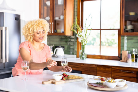 Woman in pink top arranging charcuterie on cutting board beside wine glasses in modern kitchen. Modern, culinary, stylish, hospitality, home, fresh, elegantの写真素材