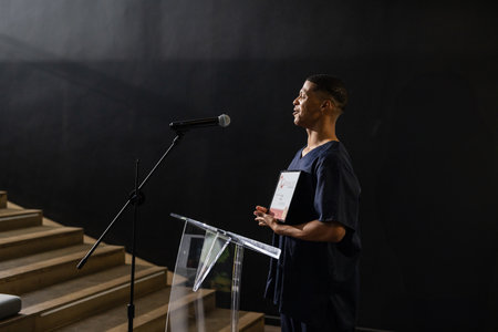 Man standing beside acrylic lectern holding certificate, using mic on stage, copy space. Public speaking, presentation, leadership, professional, conference, auditorium, motivationの写真素材