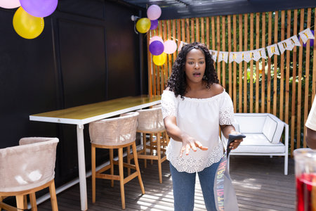 African American woman standing under pergola holding smartphone, party ribbon and birthday banner. Celebration, outdoor, joyful, leisure, festive, lifestyle, rusticの写真素材