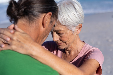 Senior woman and middle-aged man embracing and touching foreheads on sandy beach by ocean waves. Romance, intimacy, connection, seaside, serenity, outdoor, wellnessの写真素材