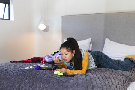 Asian woman applying makeup while lying on quilted bed surrounded by palette, brushes and clothing. Beauty, skincare, leisure, cozy, interior, femininity, lifestyleの写真素材