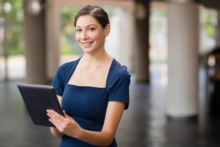 Woman holding digital tablet and smiling toward camera in spacious lobby wearing navy-blue dress. Professional, corporate, modern, architecture, interior, elegance, technologyの写真素材