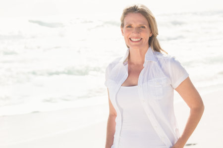 Senior woman smiling while standing on sandy shore in white shirt under bright sunlight, copy space. Seaside, leisure, outdoor, serenity, health, relaxation, minimalismの写真素材