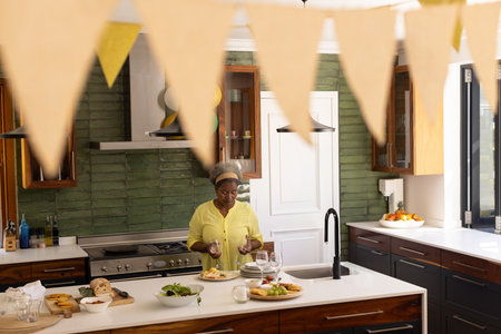 Senior African American woman plating appetizers and arranging wine glasses on island in kitchen....