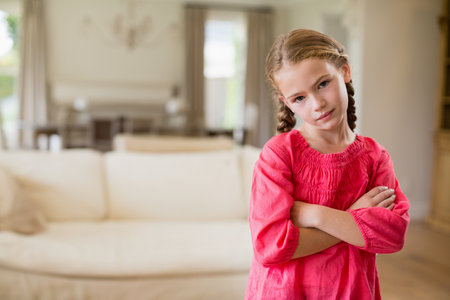 Female child standing with arms crossed in living room by sofa and chandelier, copy space. Coziness,...