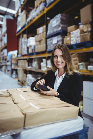 Woman in business attire standing at large warehouse scanning brown packages with handheld scanner. Logistics, inventory, industrial, commercial, organization, efficiency, technologyの写真素材