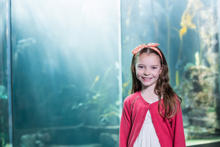 Female child wearing white dress smiling at aquarium through glass with fish and kelp, copy space. Aquarium, exploration, wonder, innocence, marine, serenity, discoveryの写真素材