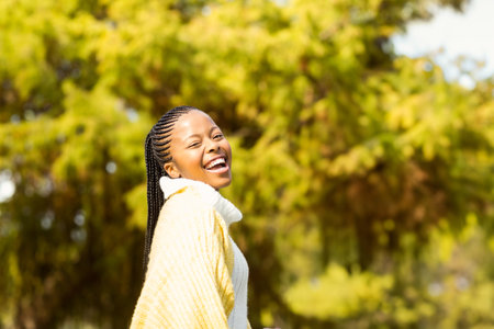 African American woman laughing while looking over shoulder in sunny park wearing sweater and shawl. Portrait, lifestyle, vibrant, natural, joyful, scenic, fashionの写真素材