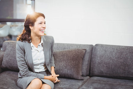 Businesswoman sitting on grey sofa and leaning forward in office lounge with sunlight streaming. Professional, collaboration, modern, corporate, workspace, communication, approachableの写真素材