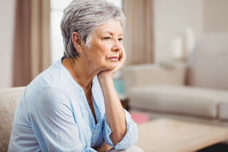 Senior woman sitting on sofa supporting cheek, gazing at coffee table at home, copy space. Retirement, relaxation, cozy, tranquility, elderly, introspection, warmの写真素材