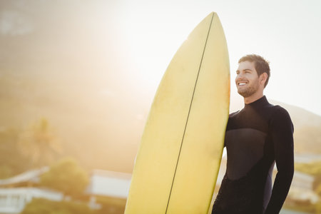 Male surfer standing on shoreline at sunset holding yellow surfboard in black wetsuit, copy space. Adventure, leisure, coast, horizon, sunlight, relaxation, sportの写真素材