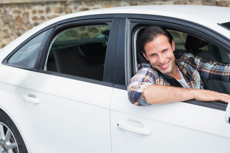Mid adult man leaning out driver side window, smiling in driveway by white sedan, copy space....