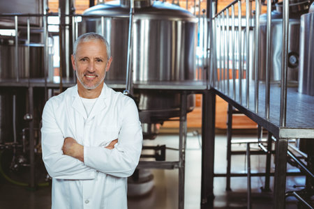 Senior man standing with crossed arms in brewery wearing lab coat beside steel fermentation tanks....