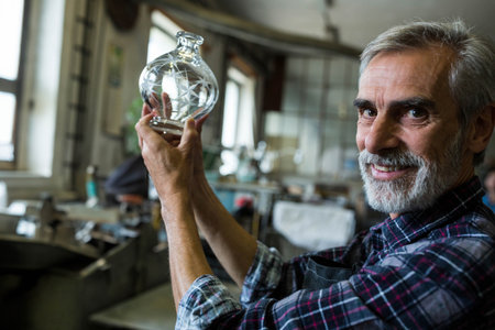 Senior craftsman holding cut glass vase up toward light in workshop with workbenches, copy space....