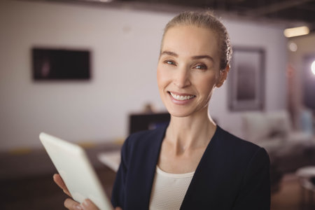 Female professional wearing blazer standing in office lounge holding tablet and smiling at camera. Corporate, workspace, modern, business, collaboration, technology, professionalの写真素材