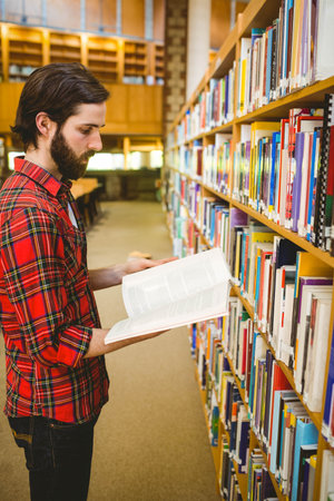 Adult male standing in library aisle between wooden bookshelves while reading open book, copy space....