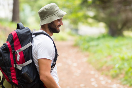 Adult male hiker standing on dirt trail with large red black backpack, bucket hat, copy space....