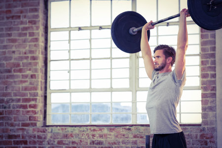 Male athlete performing overhead barbell press in studio with brick wall and window, copy space....