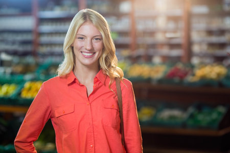 Woman smiling in red-orange shirt carrying purse strap at grocery store bins of produce. Healthy living, fresh produce, lifestyle, vibrant, wellness, shopping, organicの写真素材