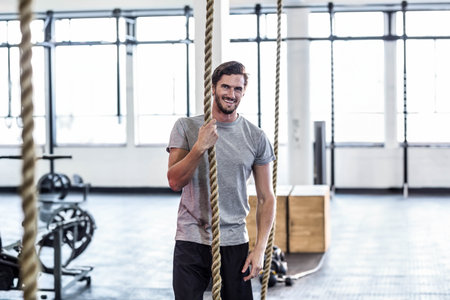 Man grasping thick climbing rope inside gym with weight racks wearing gray athletic tee and shorts....