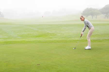 Senior woman lining up putt by flag on dew-covered green with putter and ball, copy space. Luxury,...