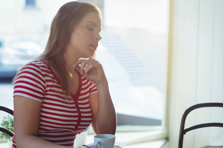 Woman sitting at cafe table by window resting chin on hand gazing over white coffee cup....