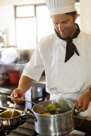 Male chef wearing uniform stirring green vegetables in steel pot on gas stove in restaurant kitchen....